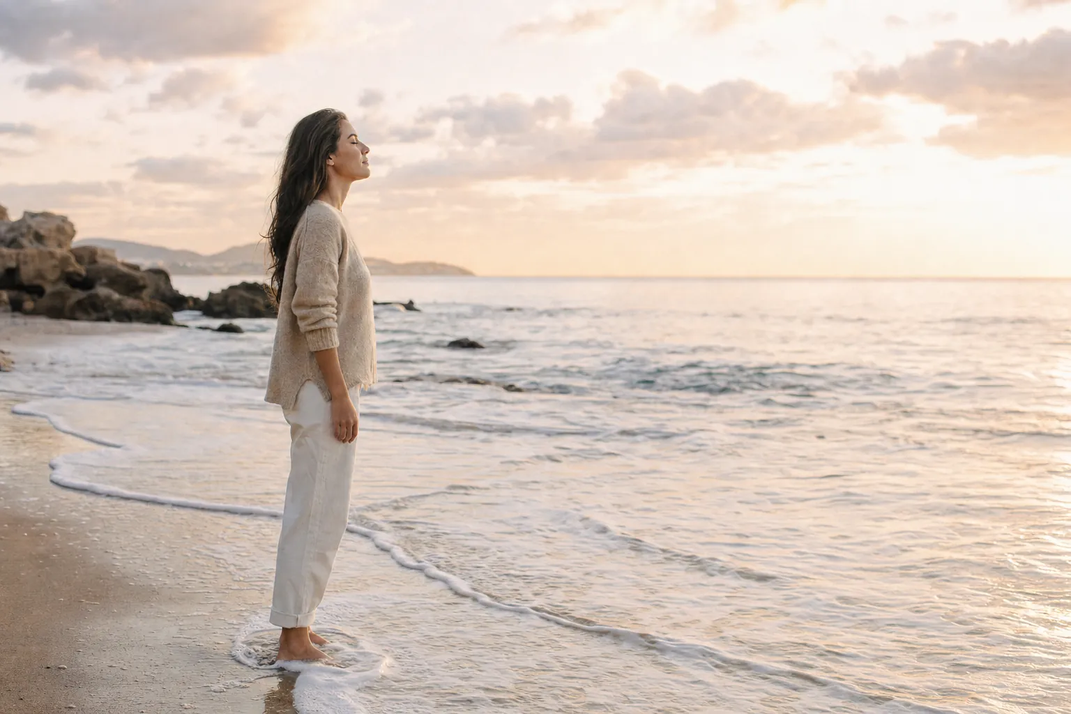 A woman pausing at the water's edge at sunrise, eyes closed, breathing slowly